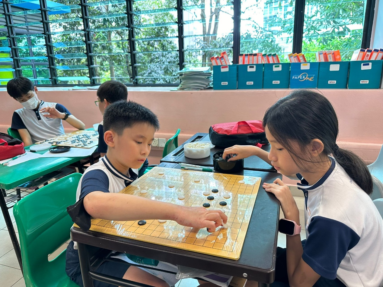 Two children in school uniforms play a game of Go with black and white stones on a grid board in a classroom.