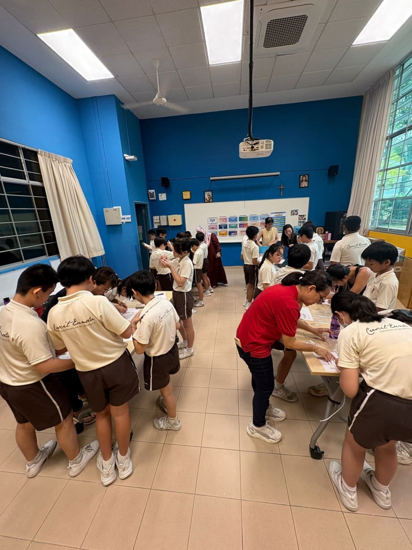 Students in matching cream and brown uniforms stand in groups writing on papers in a blue-walled classroom.