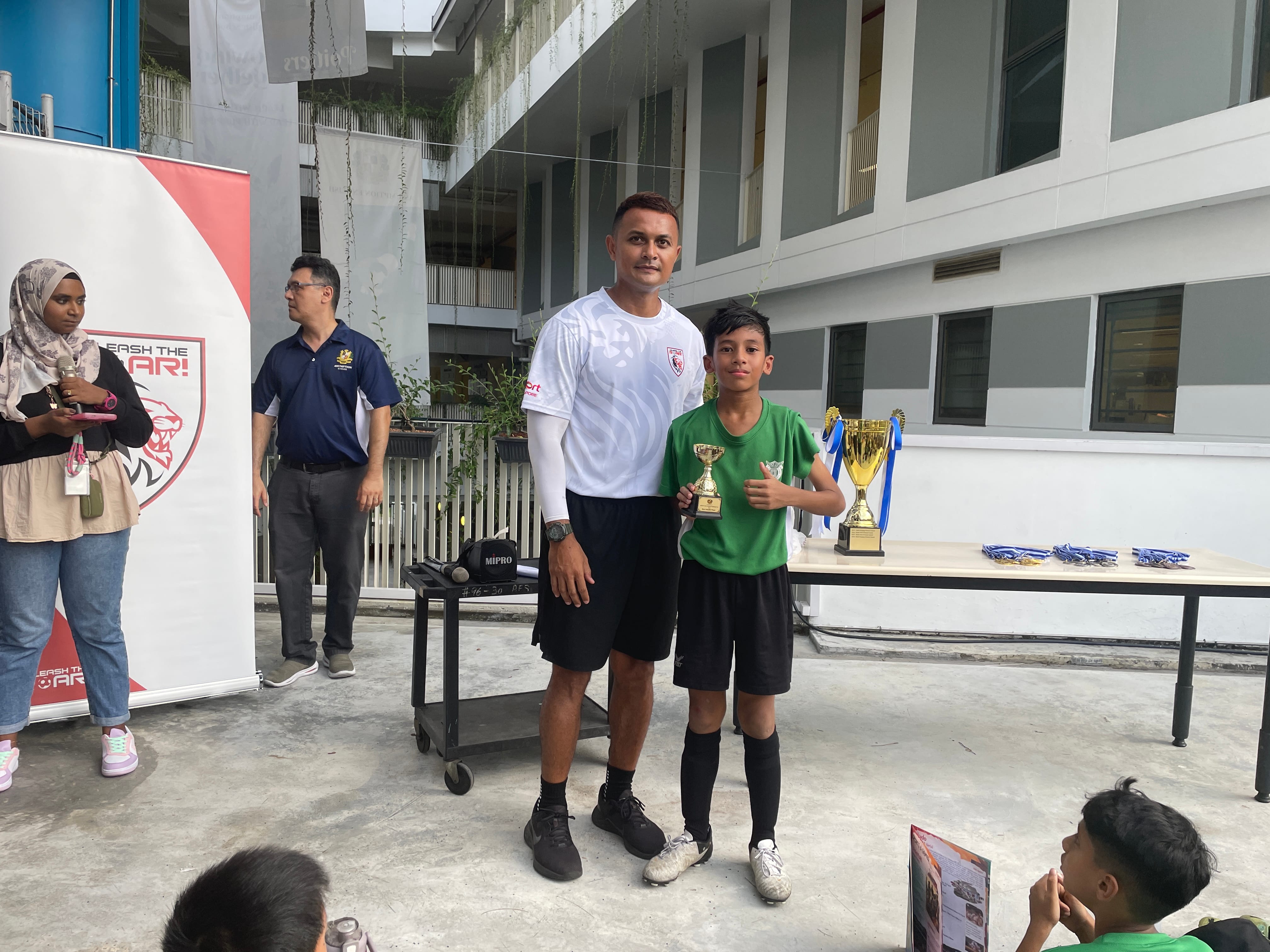 A man in a white shirt poses with a young boy holding a trophy at an outdoor sports award ceremony.