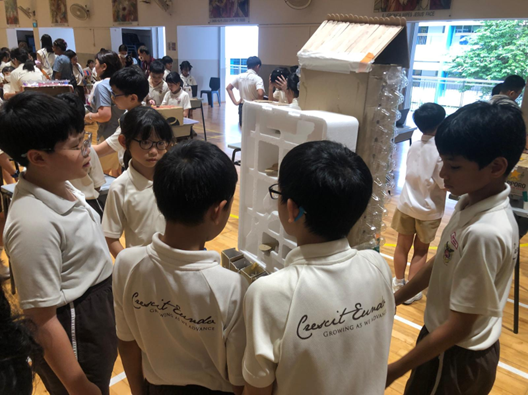 Students in matching white uniforms gather around a tall model structure built from recycled materials in a school hall.
