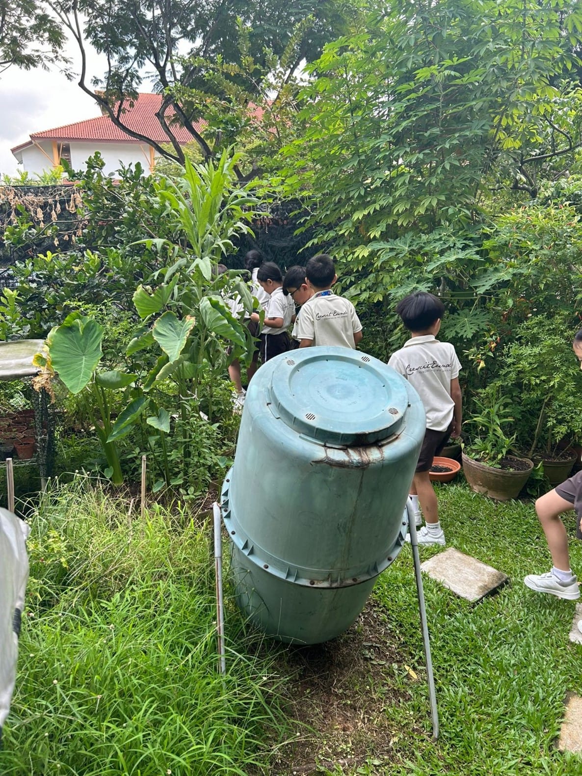 Children in school uniforms explore a lush garden with a blue compost tumbler on a stand in the foreground.