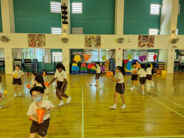 Children in school uniforms and masks do exercises holding colorful cones in an indoor gymnasium.