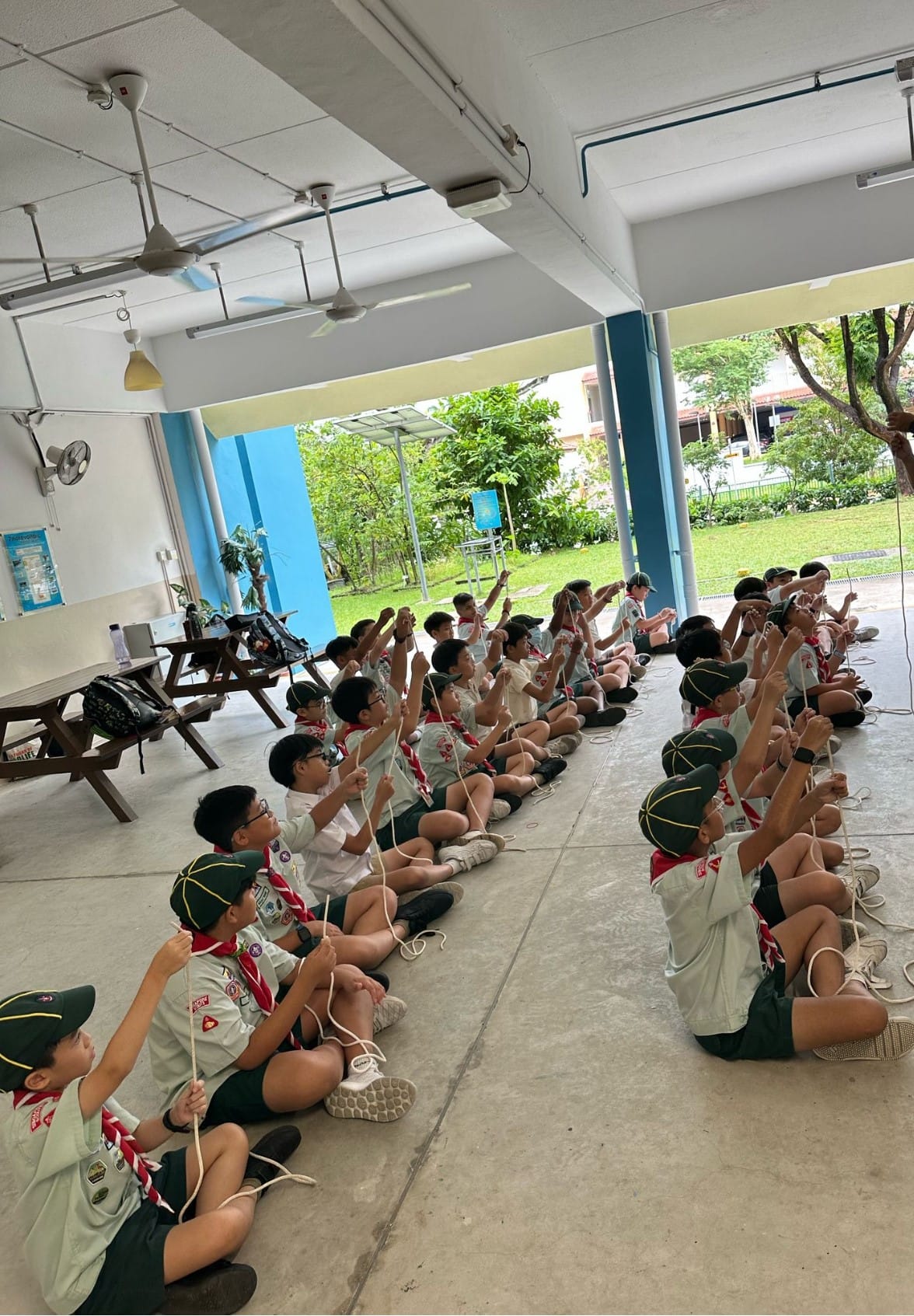 A large group of young scouts in uniform sit on a covered floor practicing rope knot tying together.