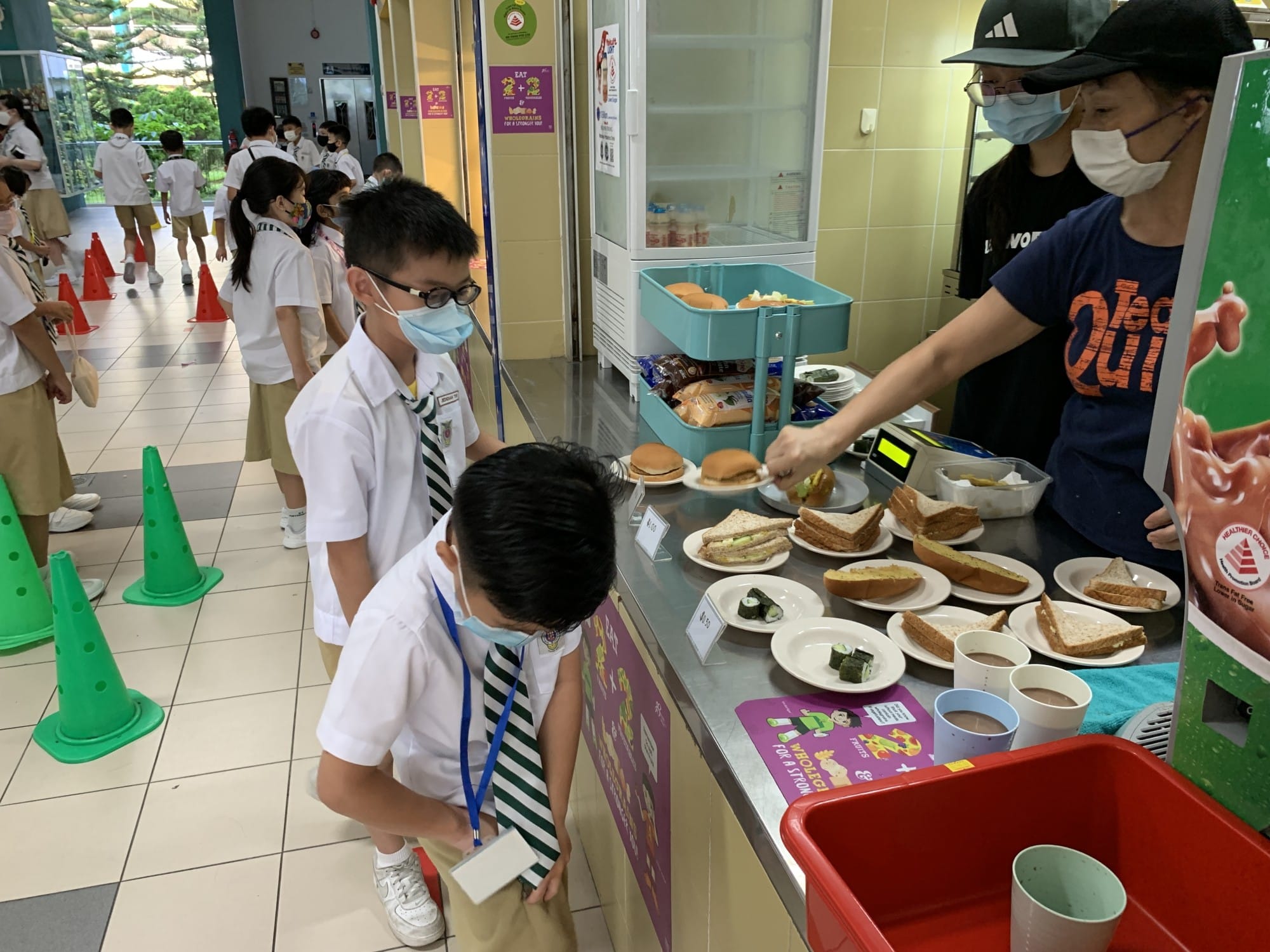 Students in school uniforms and masks queue at a canteen counter with sandwiches, burgers, and sushi served by a masked vendor.