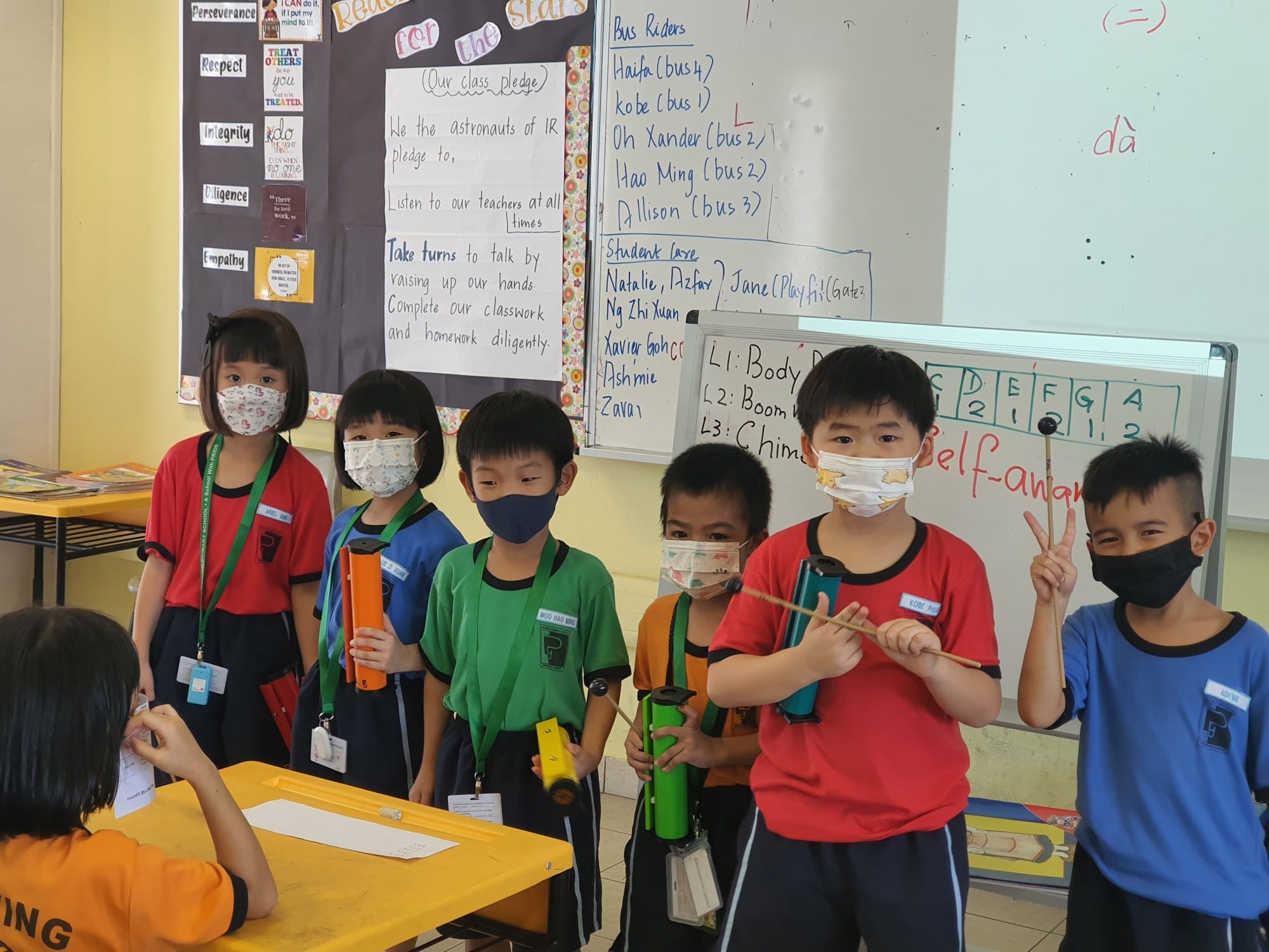 Six young masked students stand in a classroom holding colorful tube instruments, with whiteboards and posters behind them.