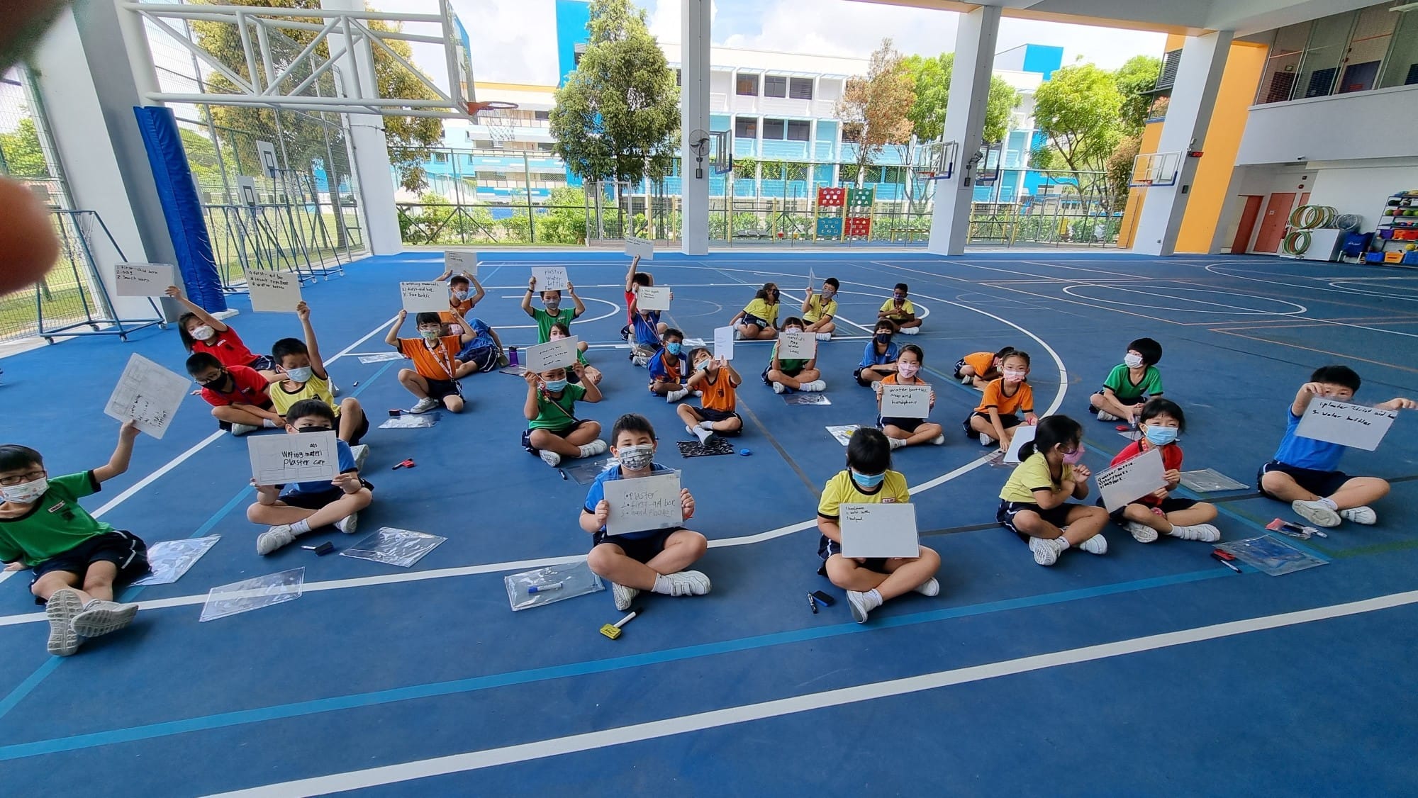 Children wearing masks sit on a blue school court holding up white boards with written notes.