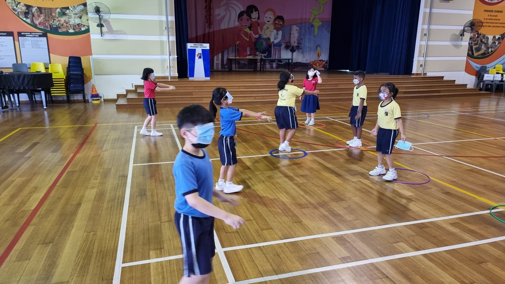 Young masked students in uniforms play with hula hoops on a school hall wooden floor near a decorated stage.