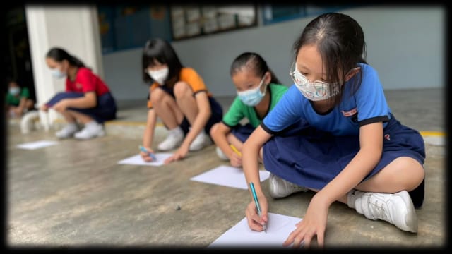 Several masked children sit on a floor writing on white paper sheets in a school corridor.