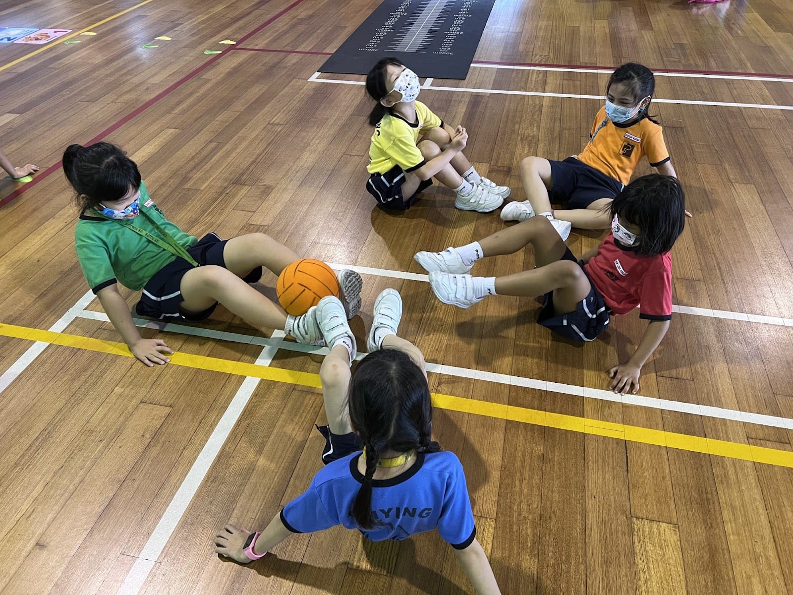 Five children wearing face masks sit in a circle on a gym floor with an orange ball between them.