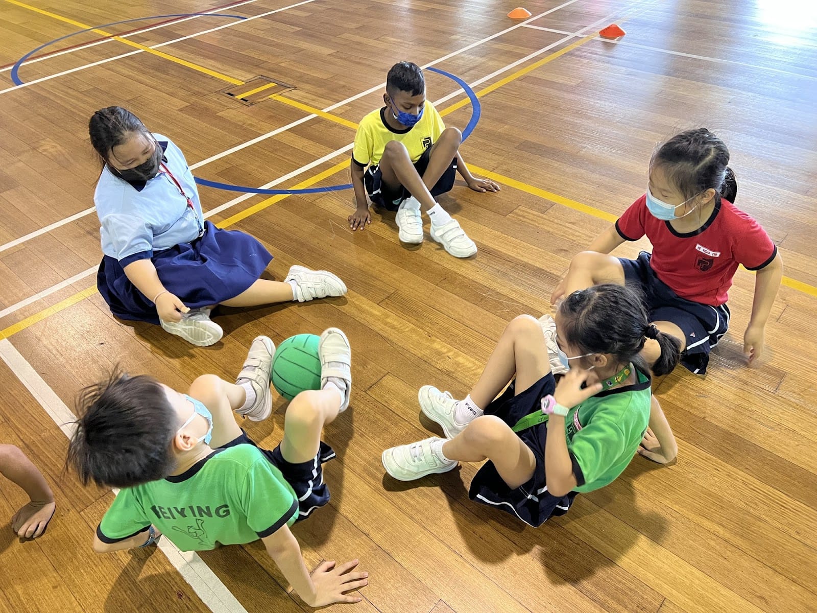 Five children wearing face masks sit in a circle on a wooden gym floor with a green ball during a PE class.