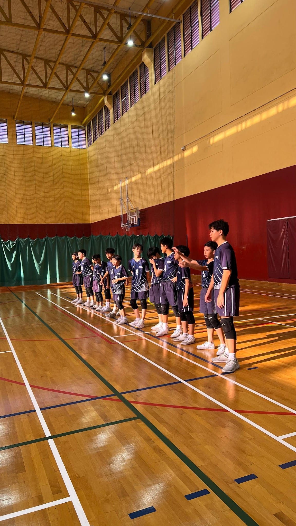 A row of young players in dark blue uniforms standing on a wooden indoor sports court while one points directions.