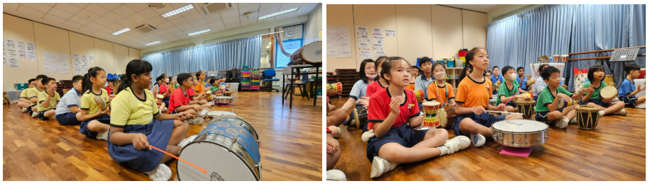 Children sit on a wooden floor in a music classroom, holding drumsticks and playing various drums.