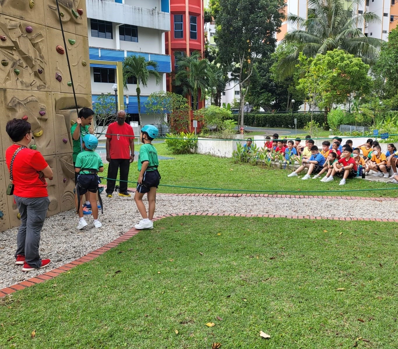 Children in helmets prepare to climb an outdoor rock wall while a large group of students watches from a grassy area.