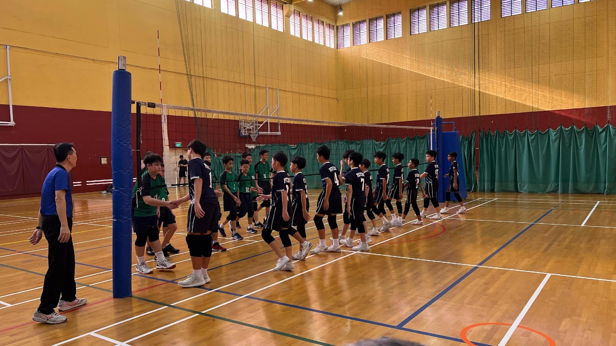 Two youth volleyball teams in black and green uniforms shake hands across the net in an indoor gymnasium.