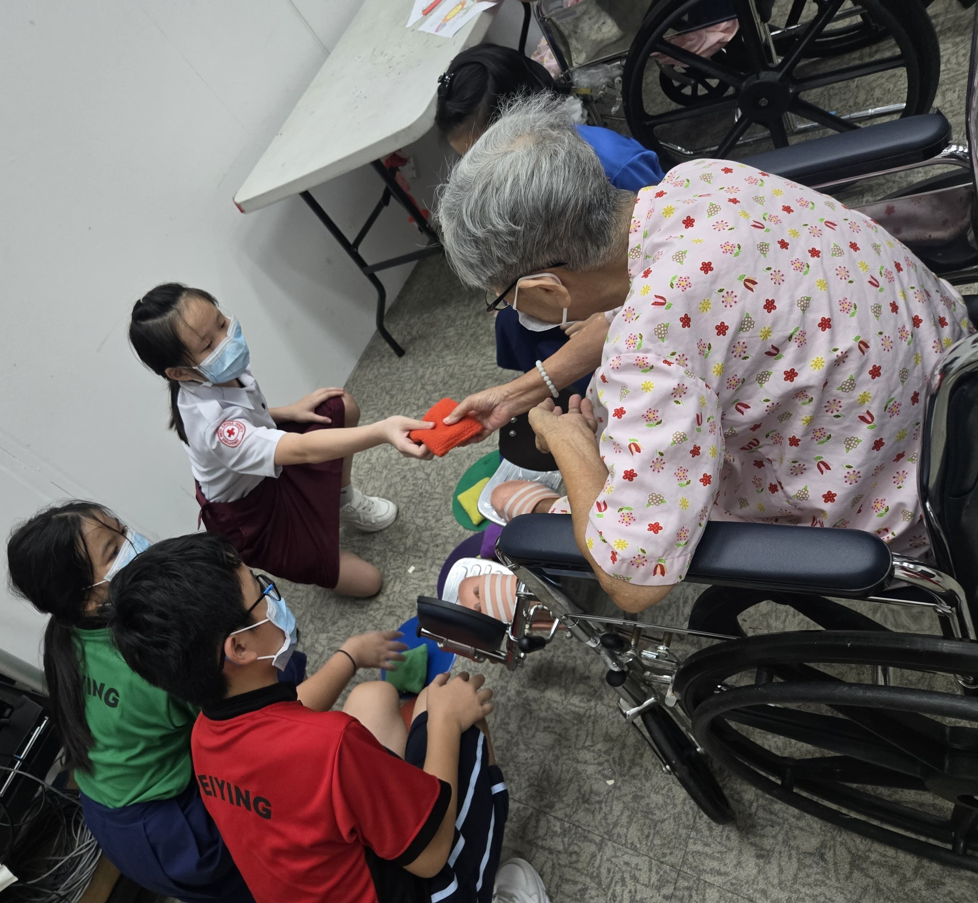 A primary school student in uniform hands a small orange bag to an elderly person sitting in a wheelchair.