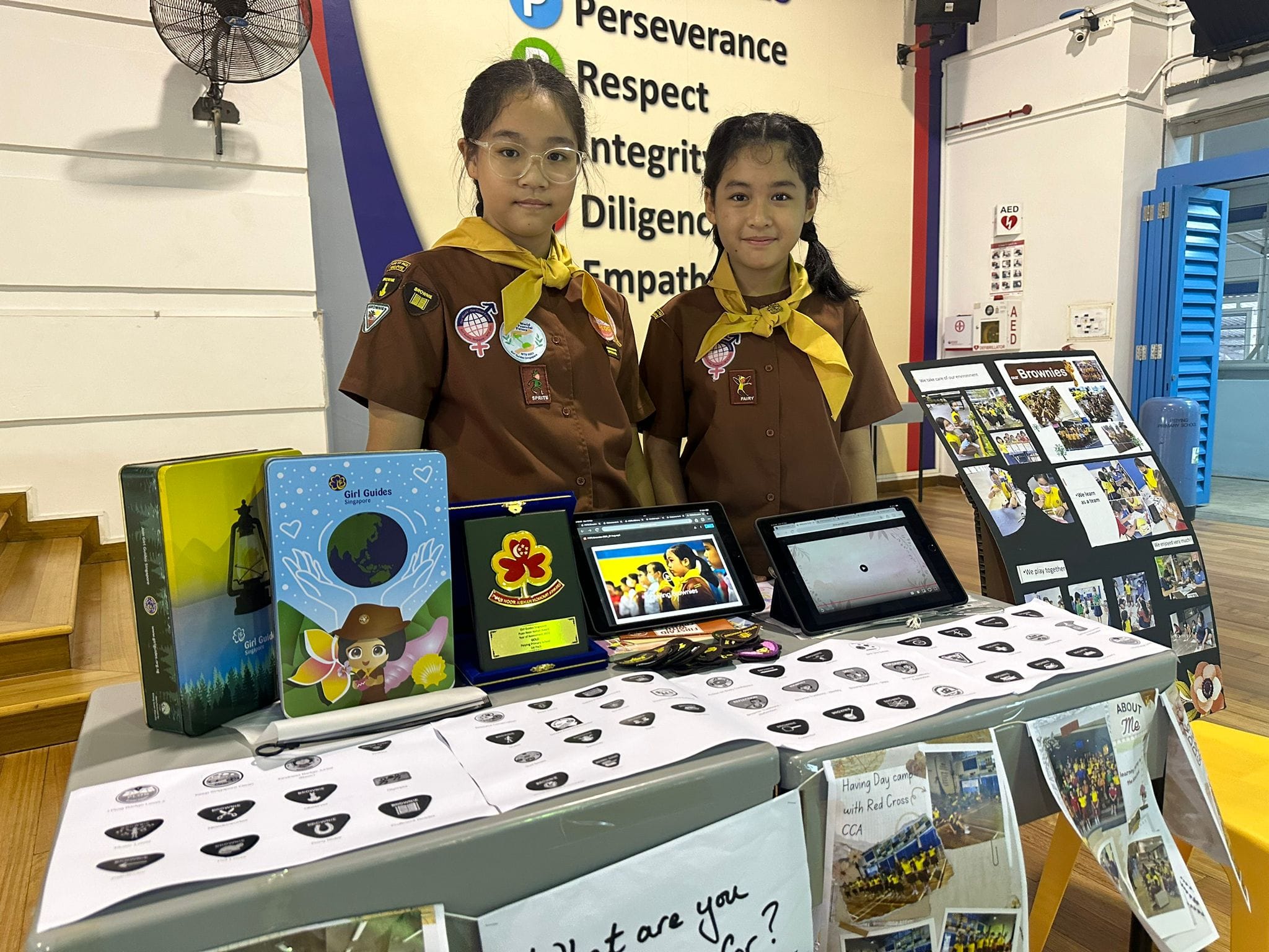 Two Girl Guides in brown uniforms stand behind a display table with tablets, badges, and Brownies materials.