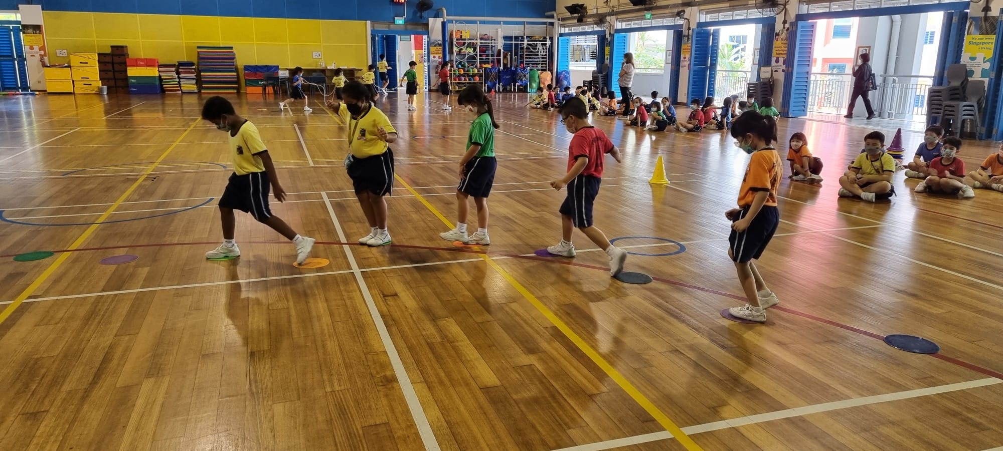 Children in school uniforms stepping on colorful floor dots in an indoor gym while others sit watching.