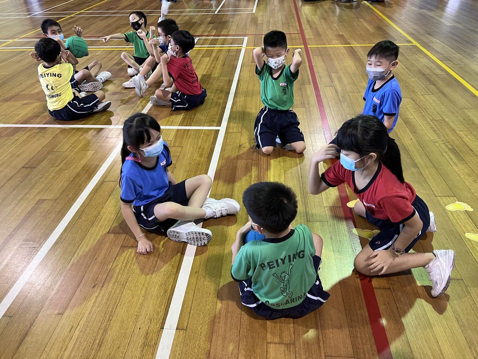 Children wearing face masks sit on a wooden gymnasium floor in colorful Peiying school sports uniforms.