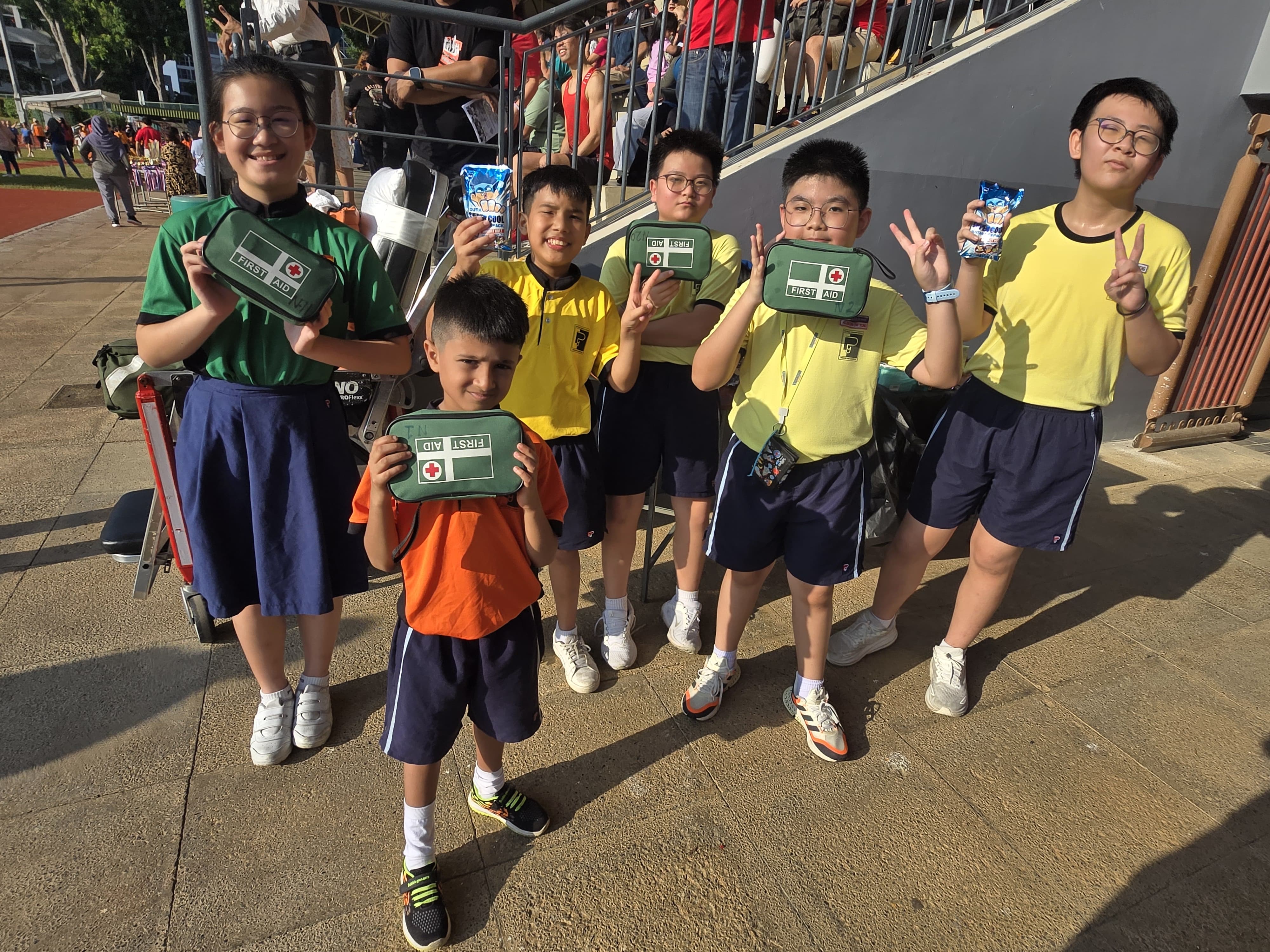 A group of primary school children in yellow and green uniforms standing together outdoors, smiling and posing with peace signs while holding green first aid kits.