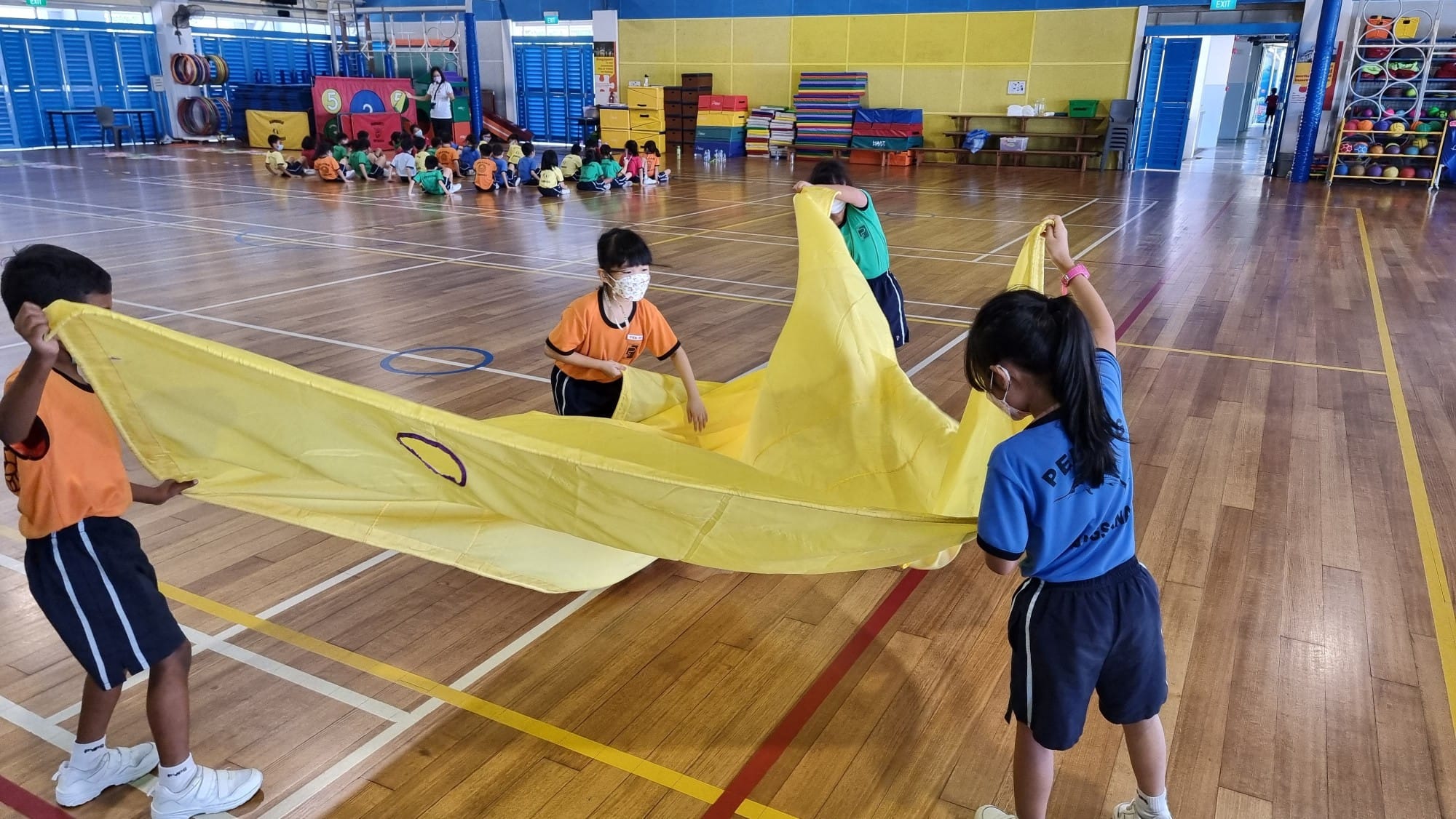 Four masked children hold up a large yellow parachute cloth in an indoor school gymnasium.