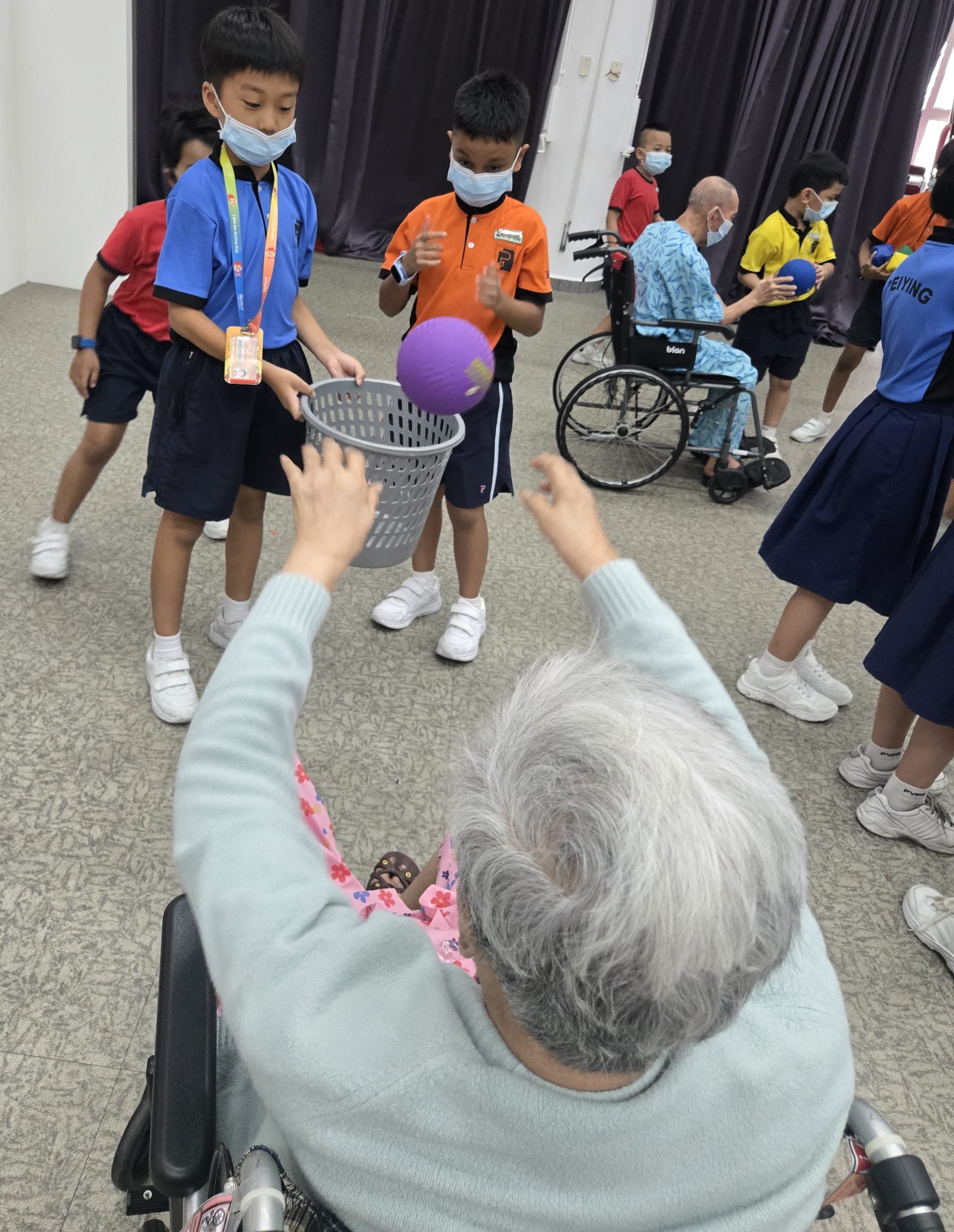 Two primary school boys playing a ball-tossing game with an elderly person in a wheelchair.