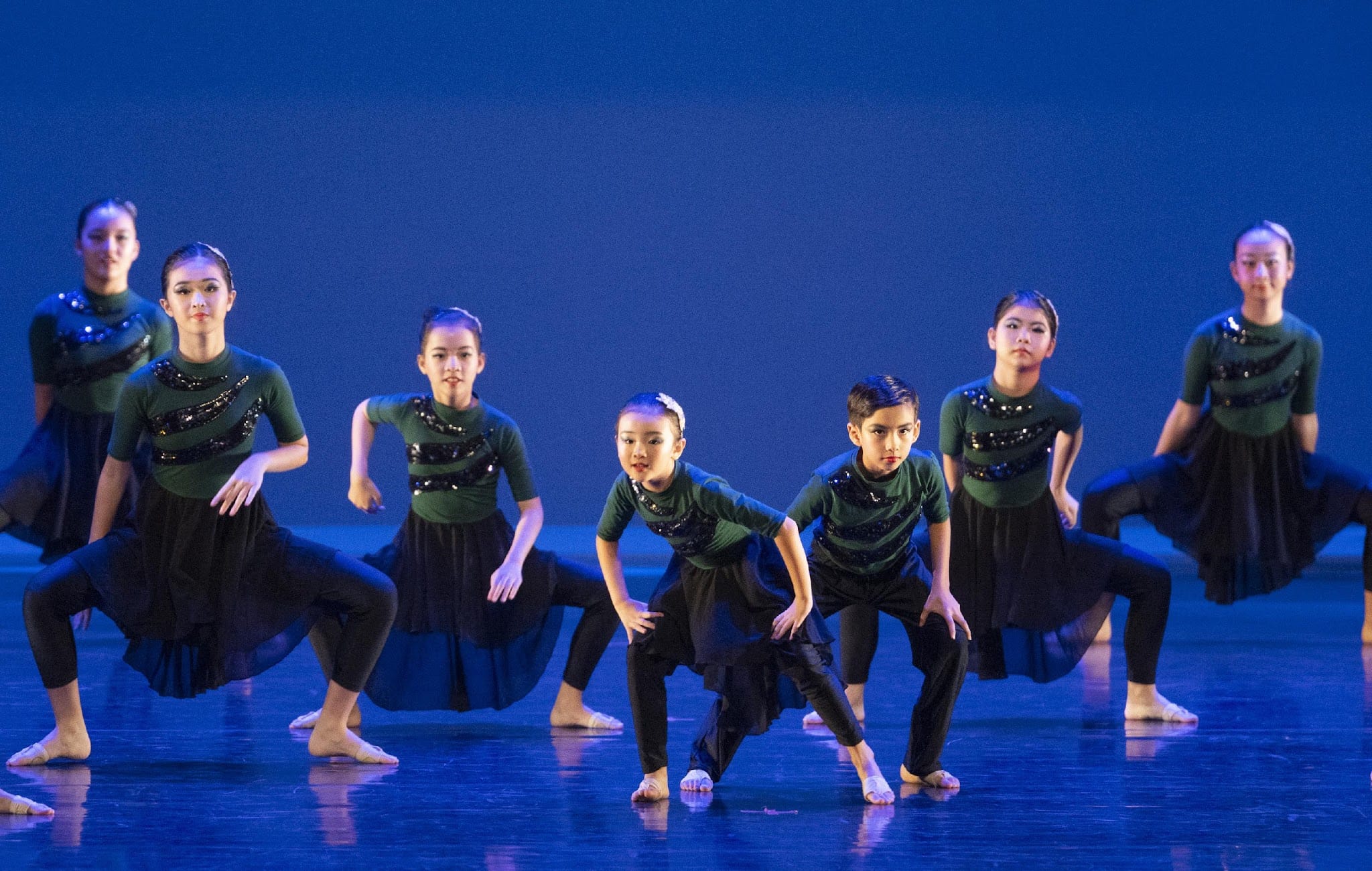 Seven young dancers perform on a blue-lit stage wearing green and black sequined costumes in synchronized poses.