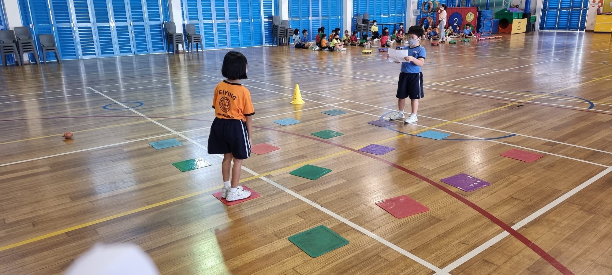 Two young children stand on colorful mats in a school gymnasium with other kids seated in the background.