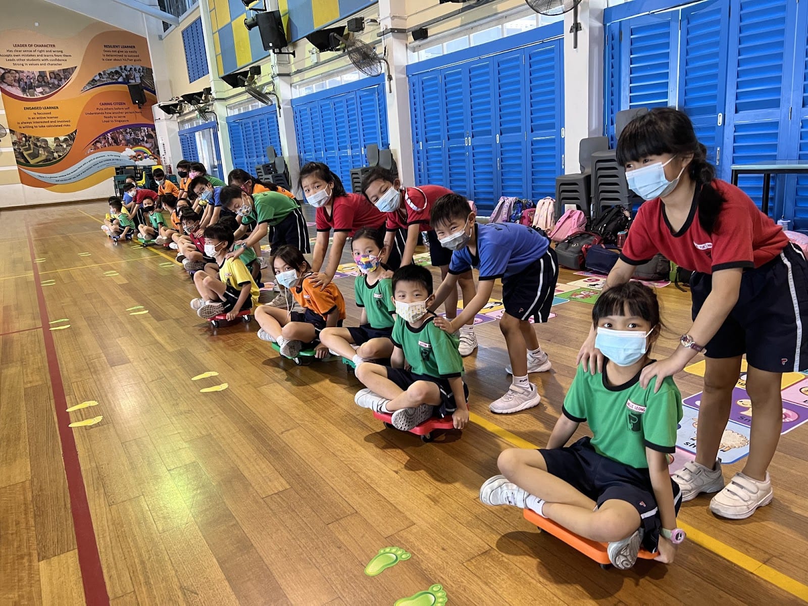 Children wearing masks sit on scooter boards in a school gym while older students push them in a line activity.