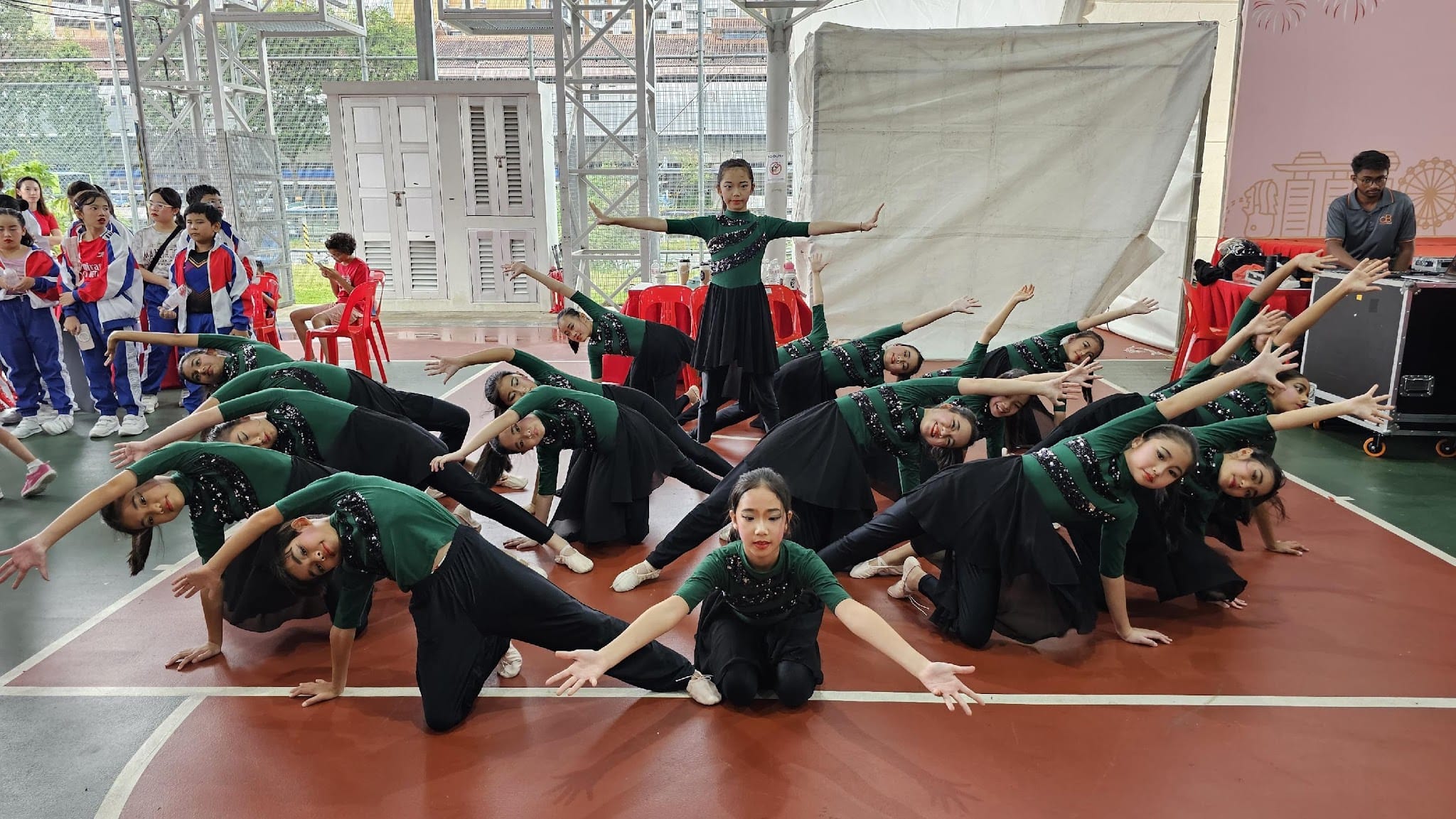 A group of young dancers in green and black outfits perform a synchronized routine on an indoor court before an audience.