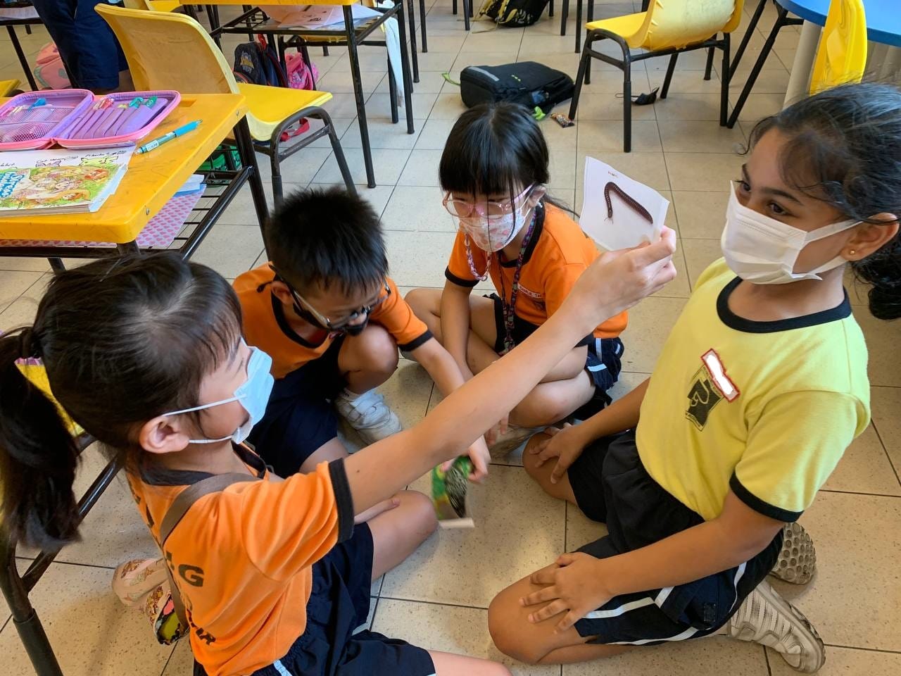 Four masked school children sit on a classroom floor, one holding a card with a worm picture.