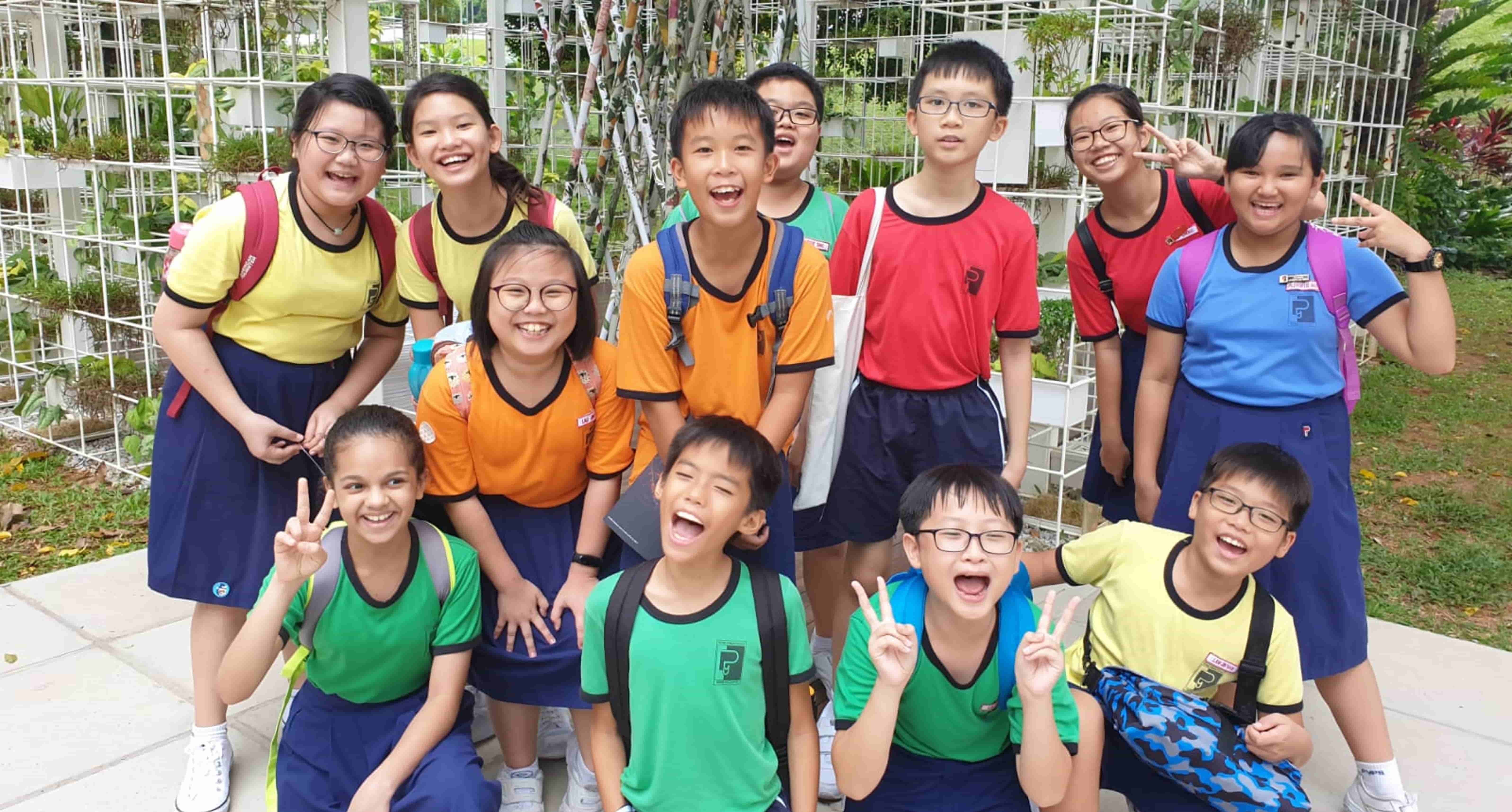 A group of smiling school children in colorful uniforms posing outdoors near a white trellis with green plants.