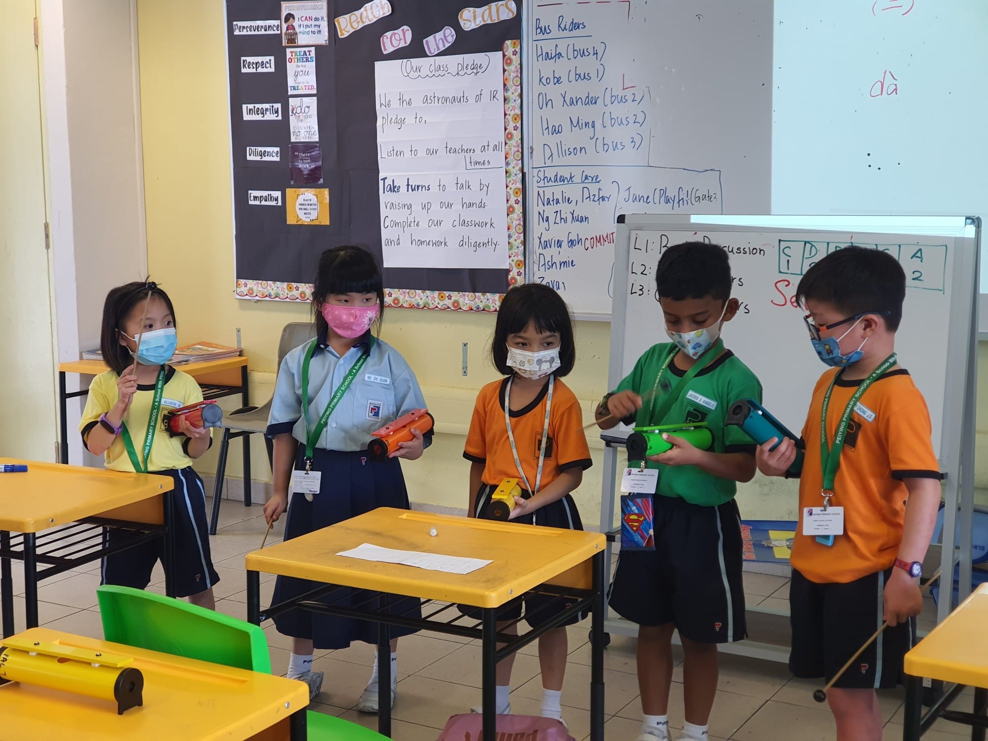 Five masked primary school children stand in a classroom holding colorful toy devices near yellow desks and a whiteboard.