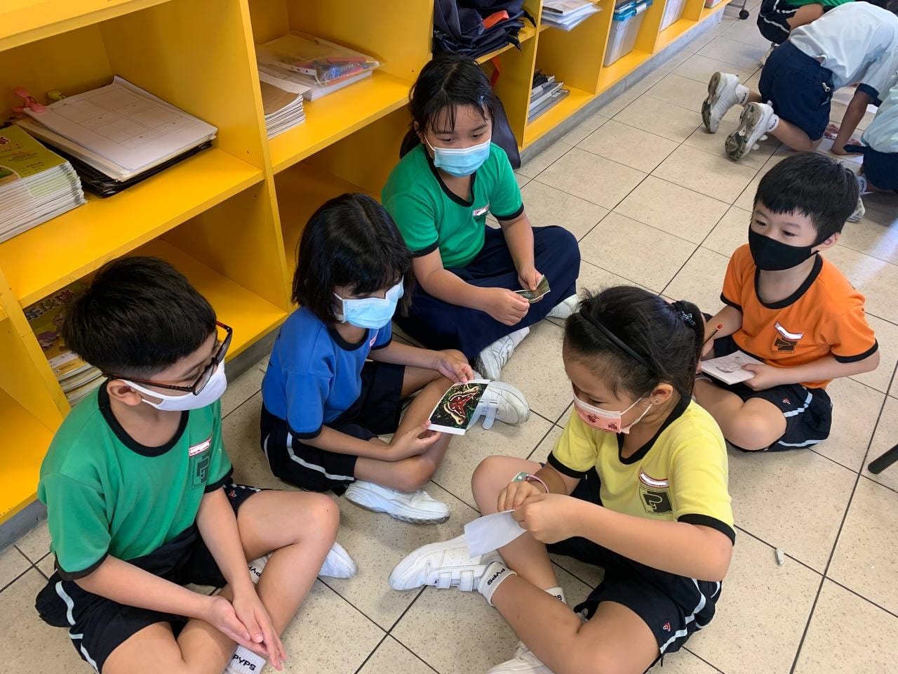 Five masked children in school uniforms sit on a tiled floor near yellow shelves, looking at cards together.