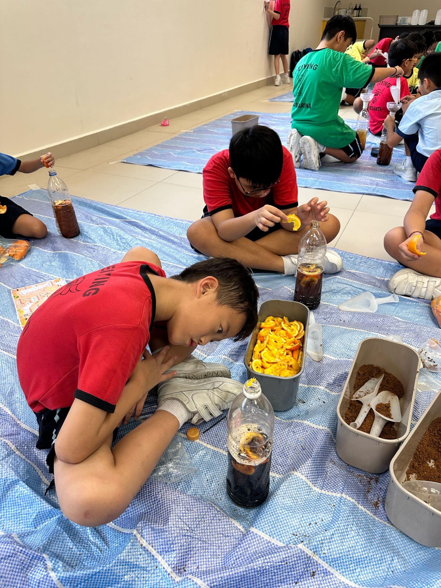 Children in red shirts sit on blue tarps on a floor, filling bottles with orange slices and dark liquid.