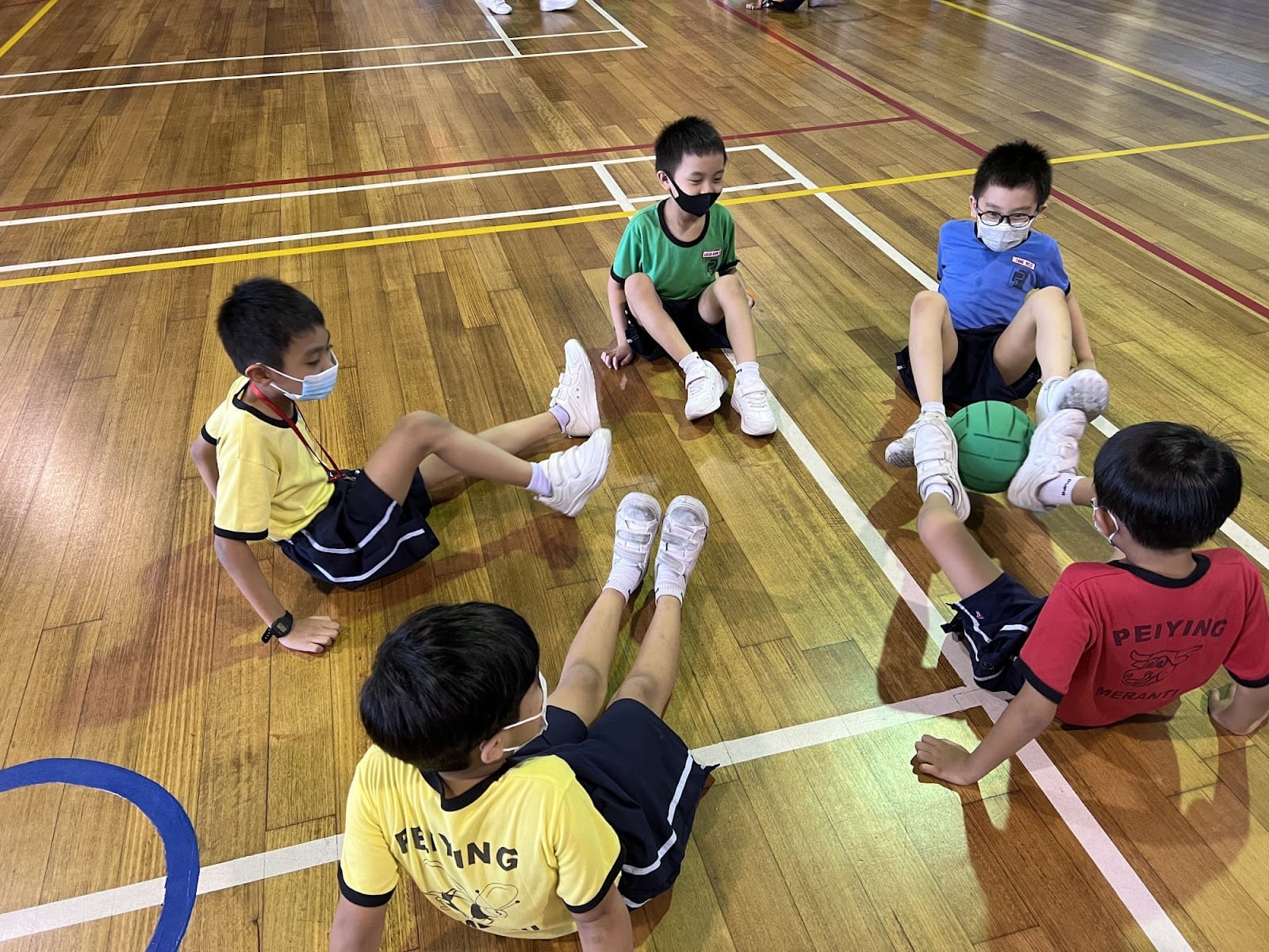 Five masked children sit on a wooden gym floor in a circle with a green ball during a school PE class.