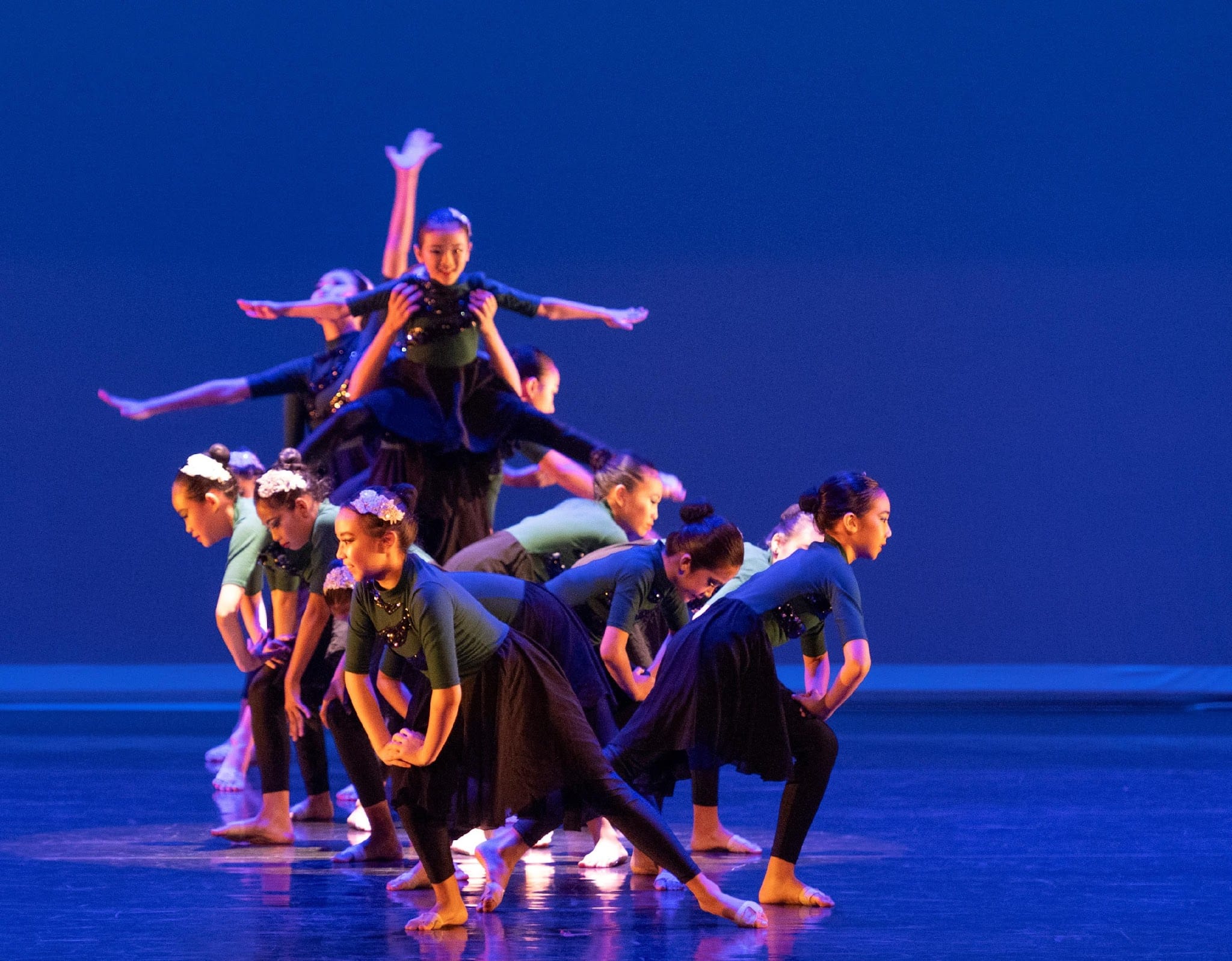 A group of young dancers in dark costumes perform a layered formation on a blue-lit stage, one lifted above the others.