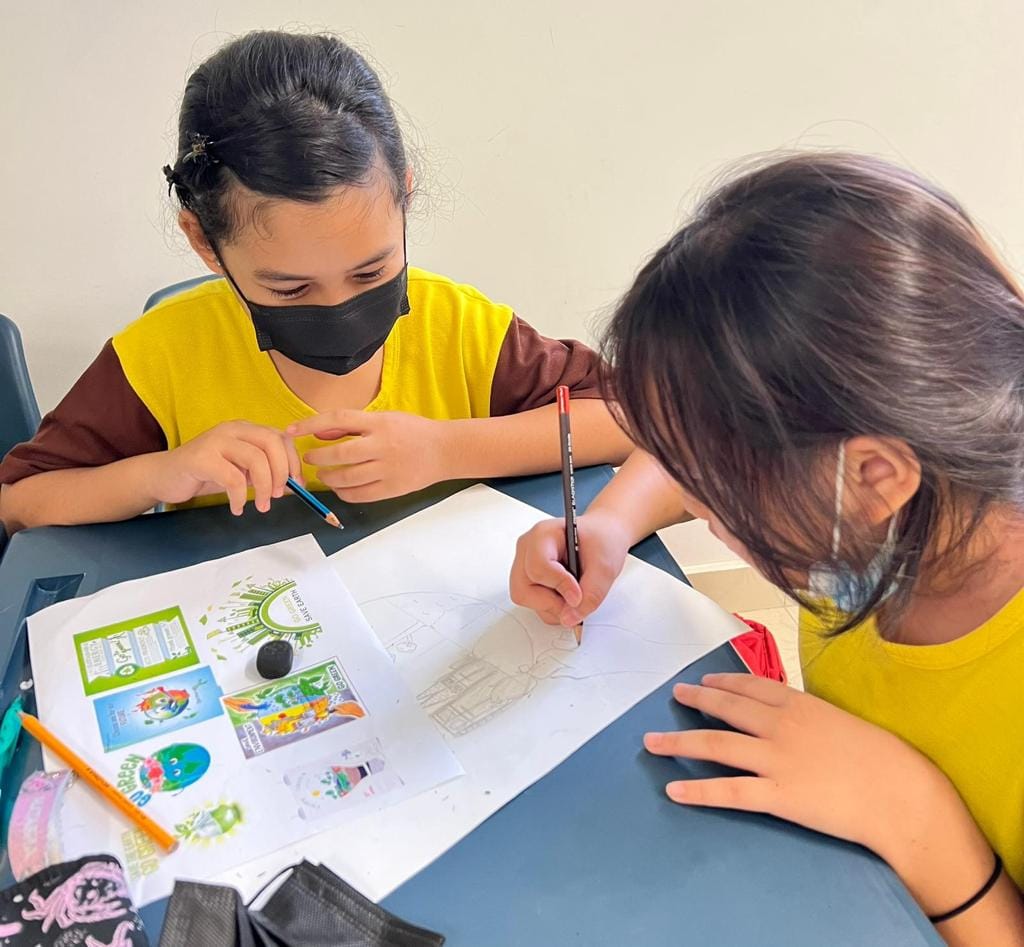 Two masked children in yellow shirts sit at a table, one drawing with a pencil on large paper with printed reference cards nearby.