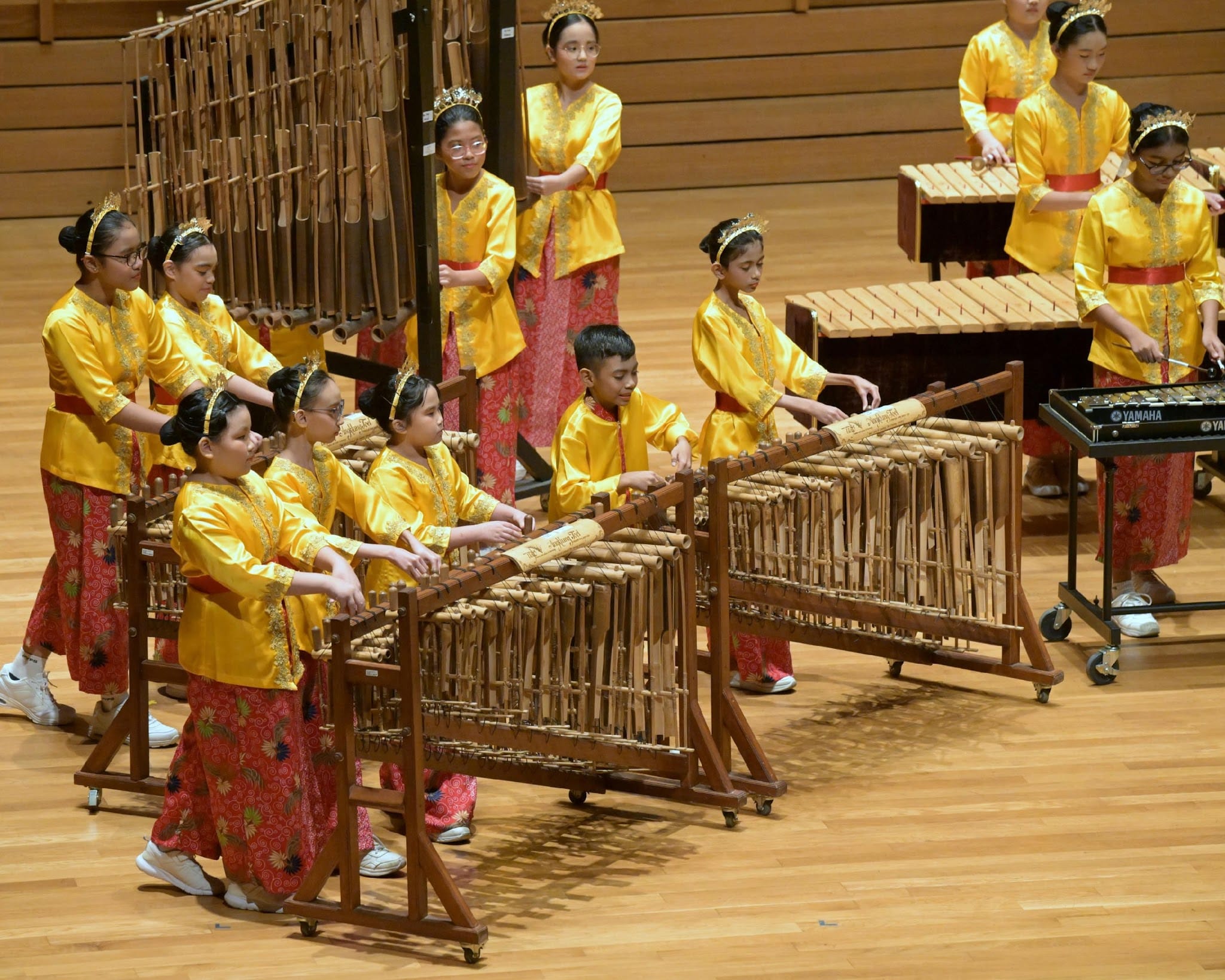 Children in gold and red traditional costumes perform on angklung bamboo instruments on a wooden stage.