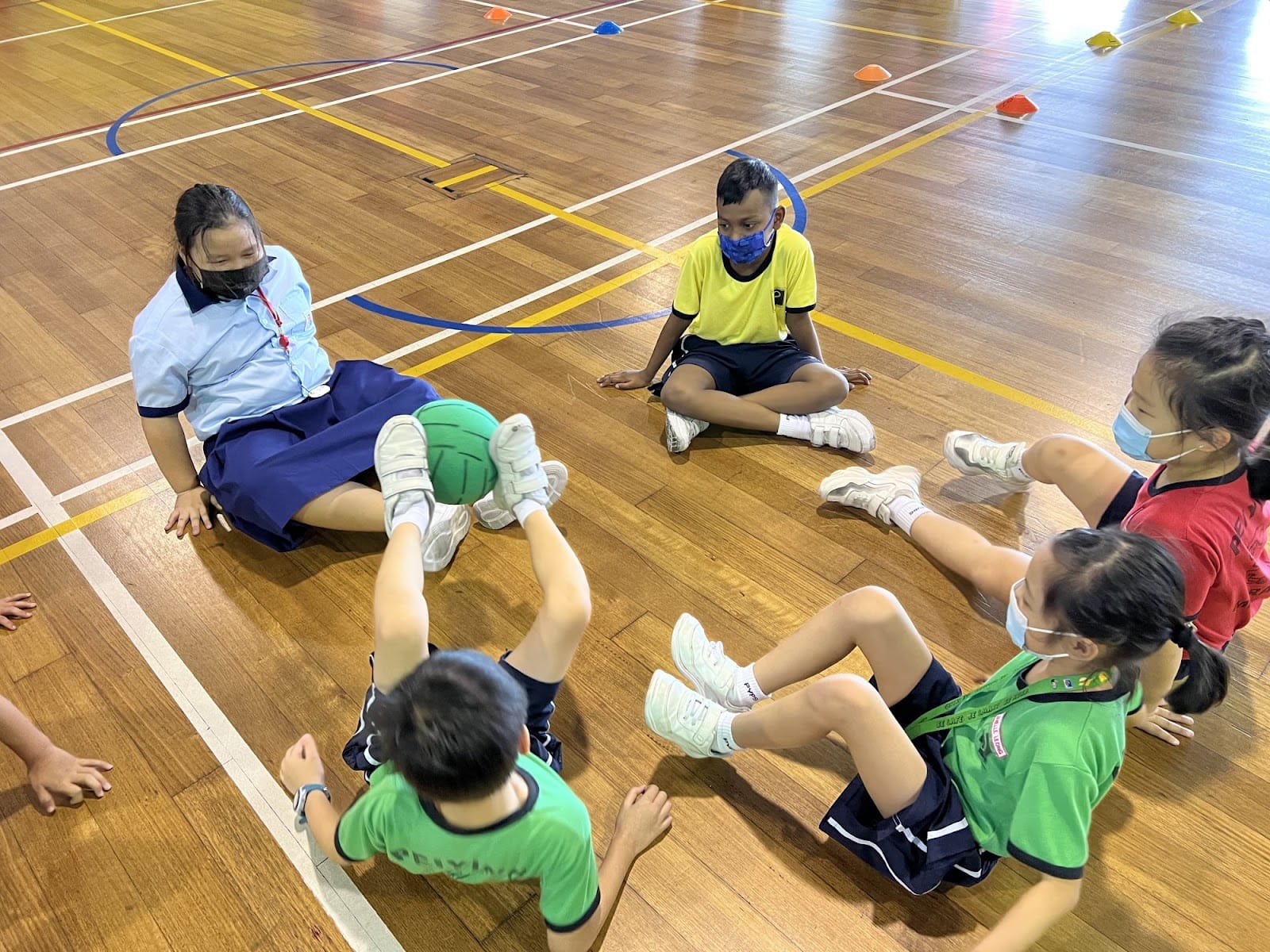 Five masked children sit on a gym floor passing a green ball with their feet during a PE activity.