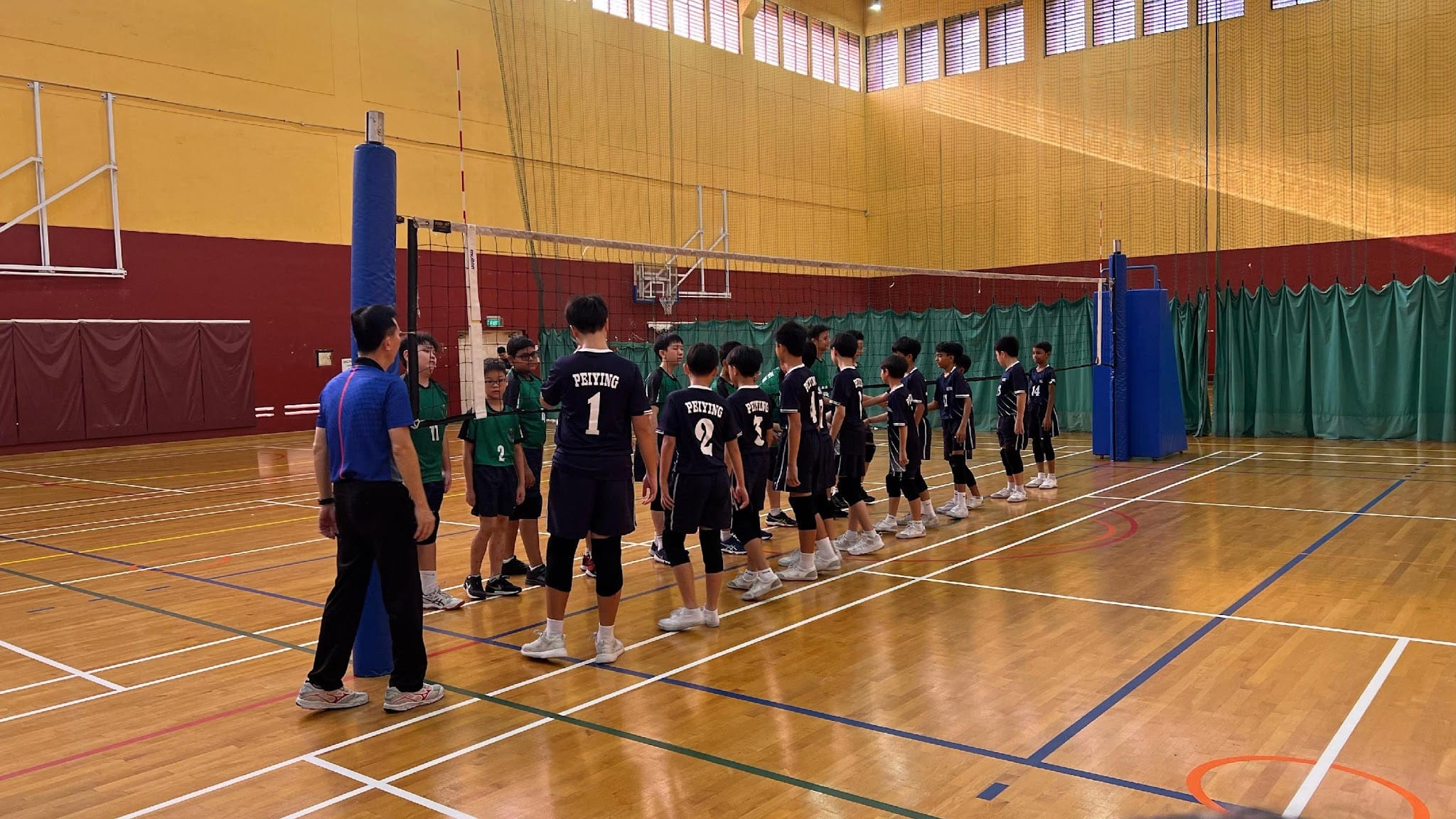 Two youth volleyball teams in green and dark jerseys line up at the net indoors as an adult referee watches.