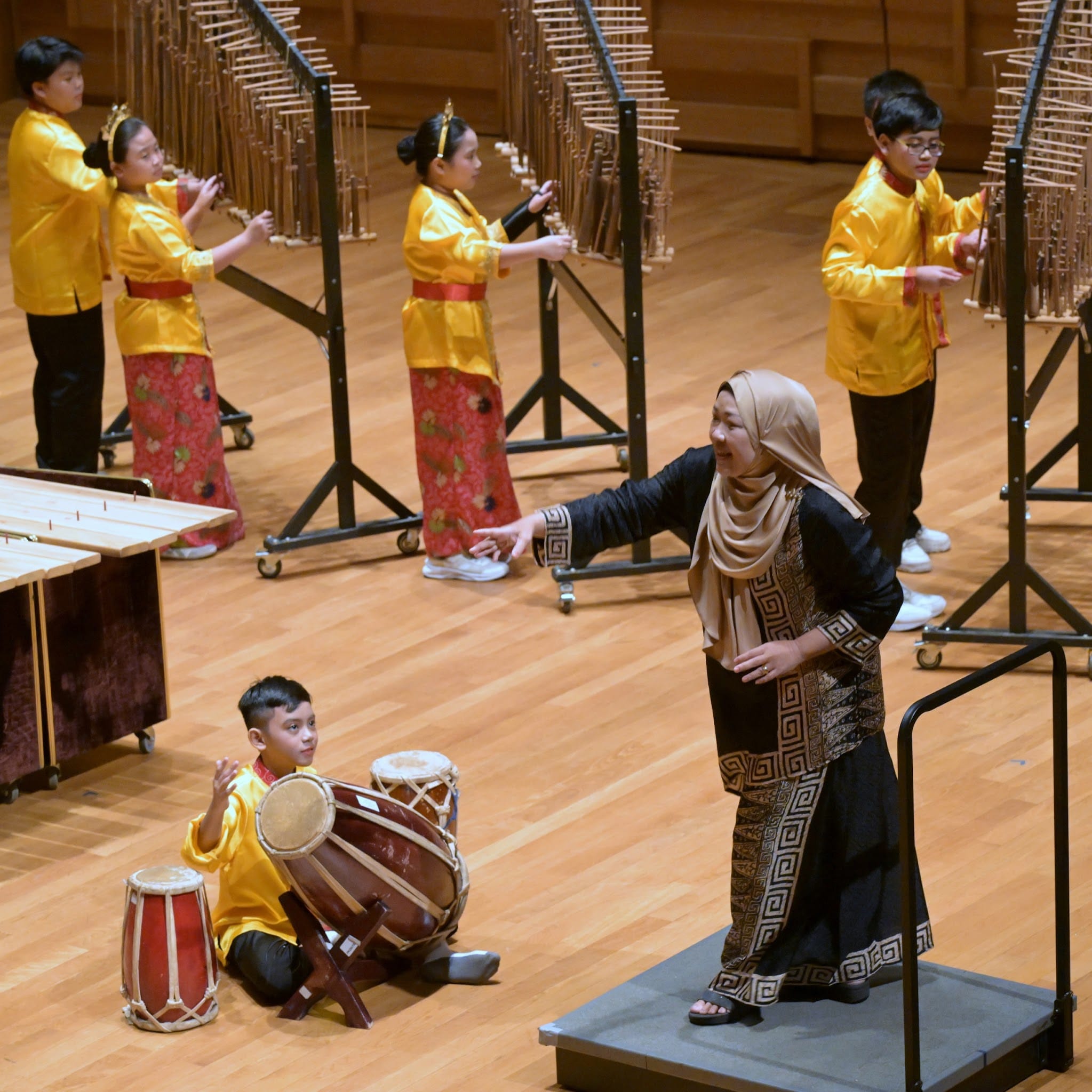 Children in gold outfits play traditional percussion instruments on a wooden stage, guided by a woman in a hijab.