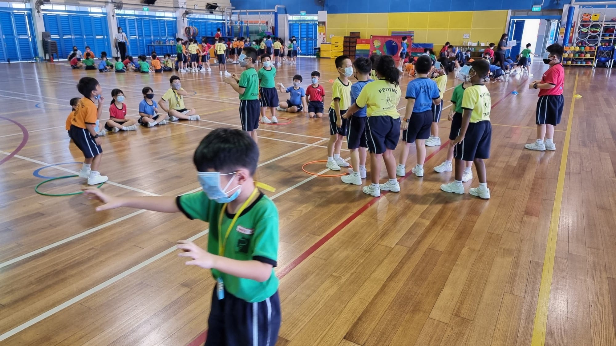 Children wearing masks and colorful shirts participate in a physical education class inside a school gymnasium.