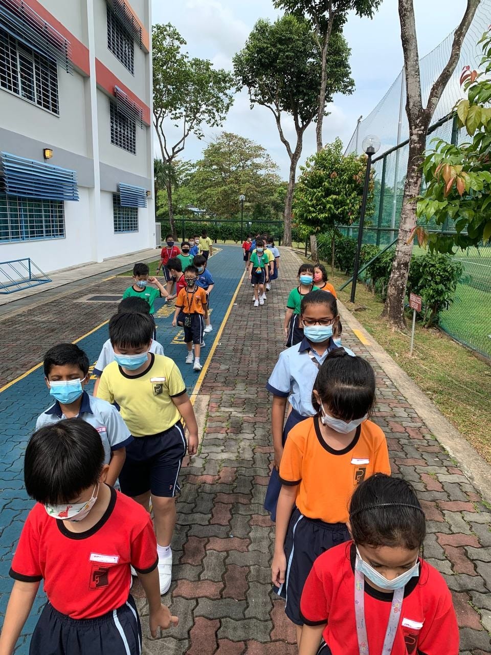 Children wearing face masks walk in two lines along a paved outdoor path beside a school building.