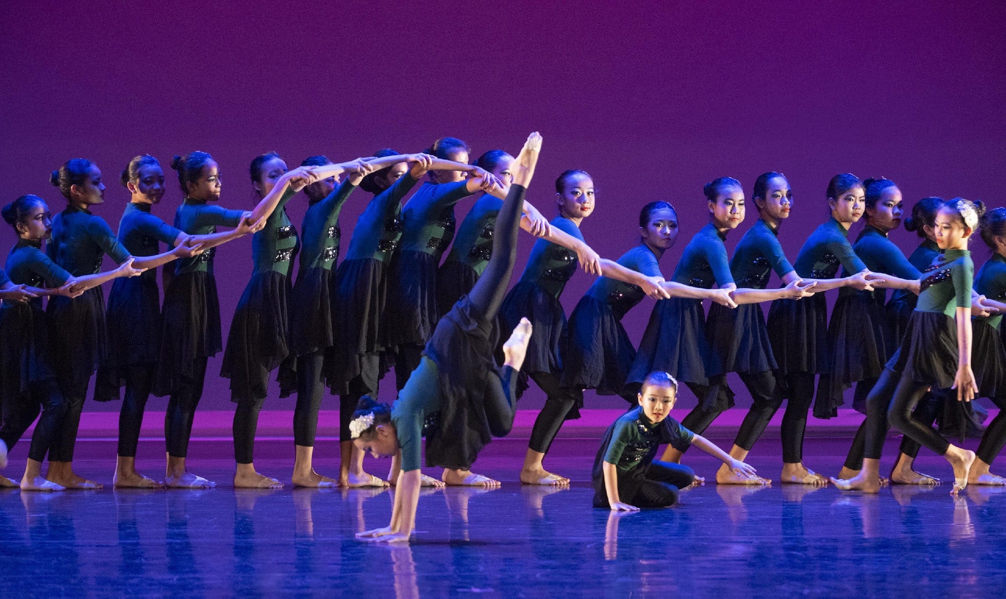 A large group of dancers in black and green costumes perform on a purple-lit stage, with one doing a handstand split.