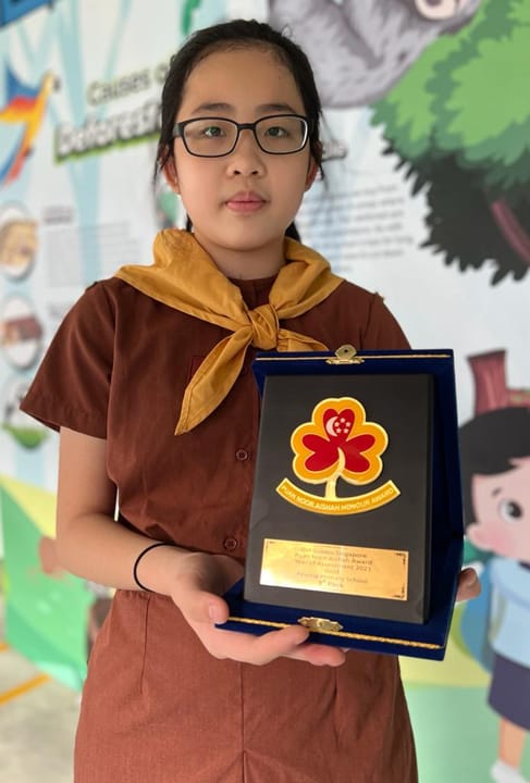 Young girl in a brown scout uniform holding a framed award plaque with a clover emblem.