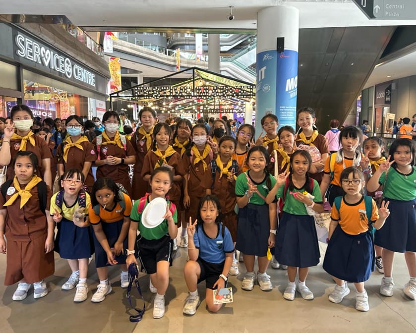 A large group of young schoolgirls in uniforms posing together inside a shopping mall, many making peace signs.
