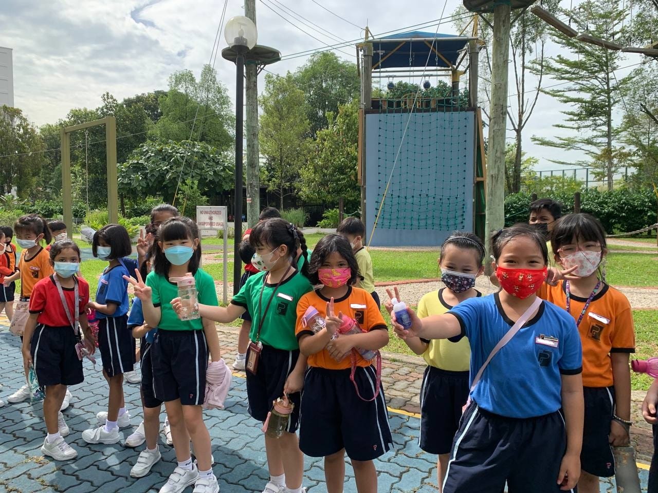 A group of young children wearing face masks and school uniforms pose with peace signs near an outdoor climbing structure.