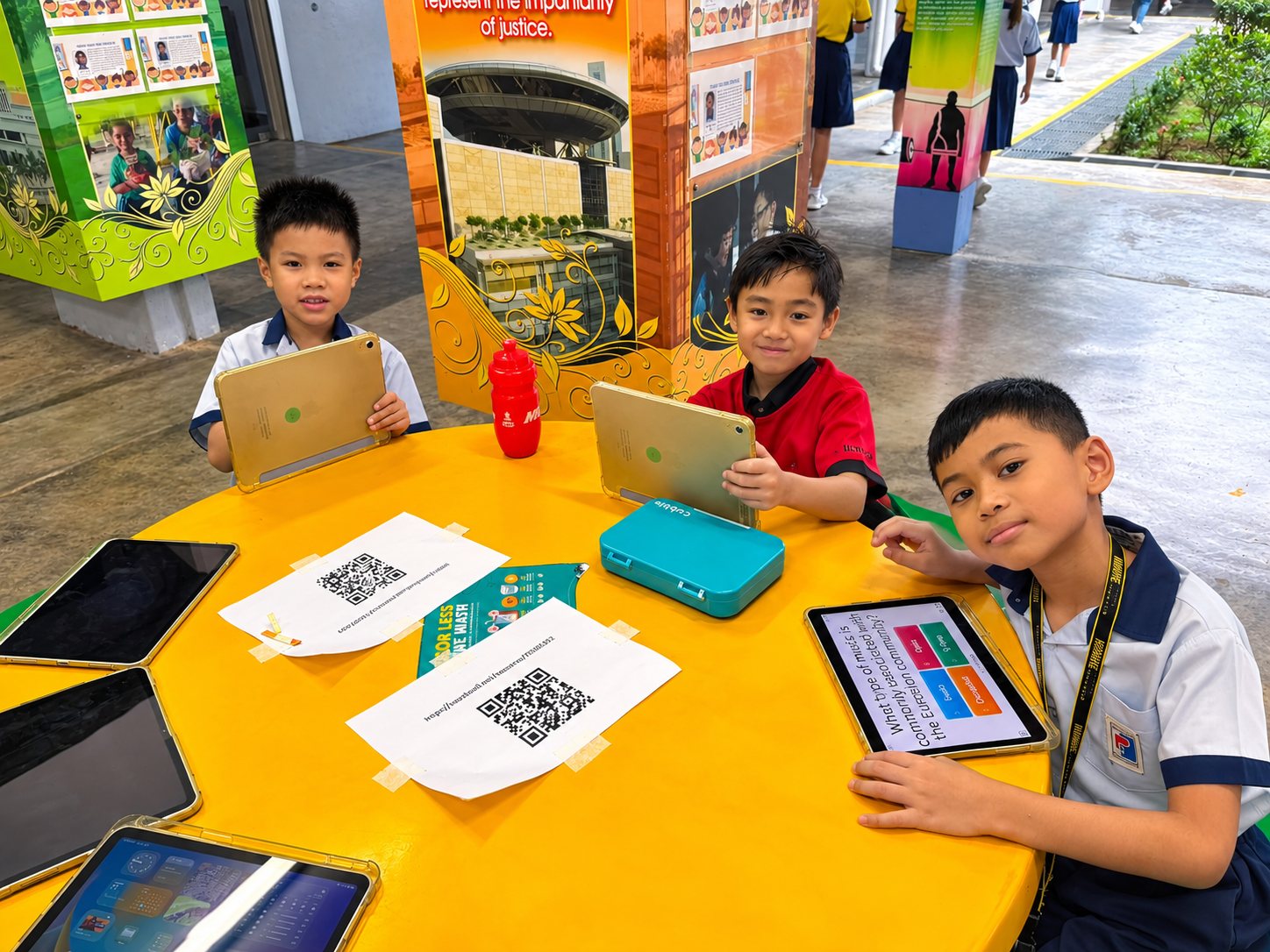 Three students sitting at a bright yellow table using tablets and printed worksheets.