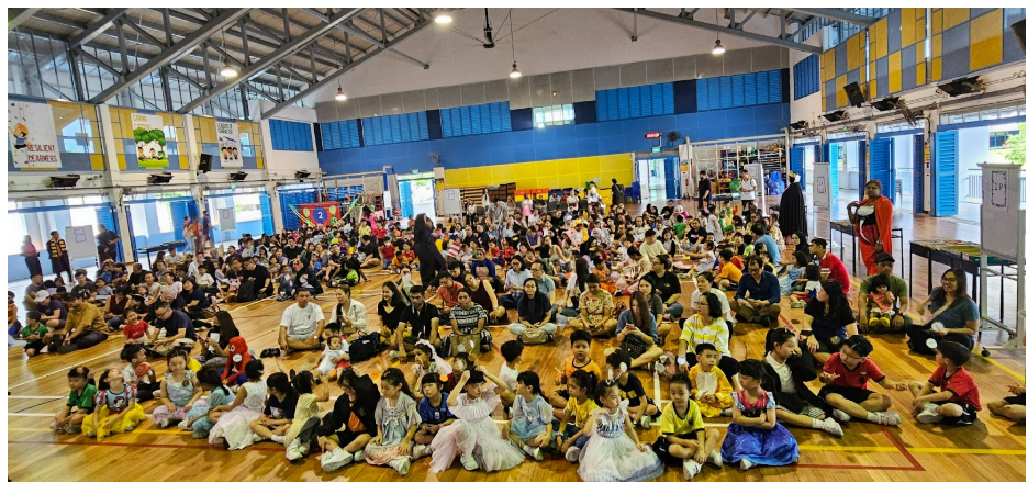 Large group of children and adults in costumes sitting on a school gymnasium floor during an event.