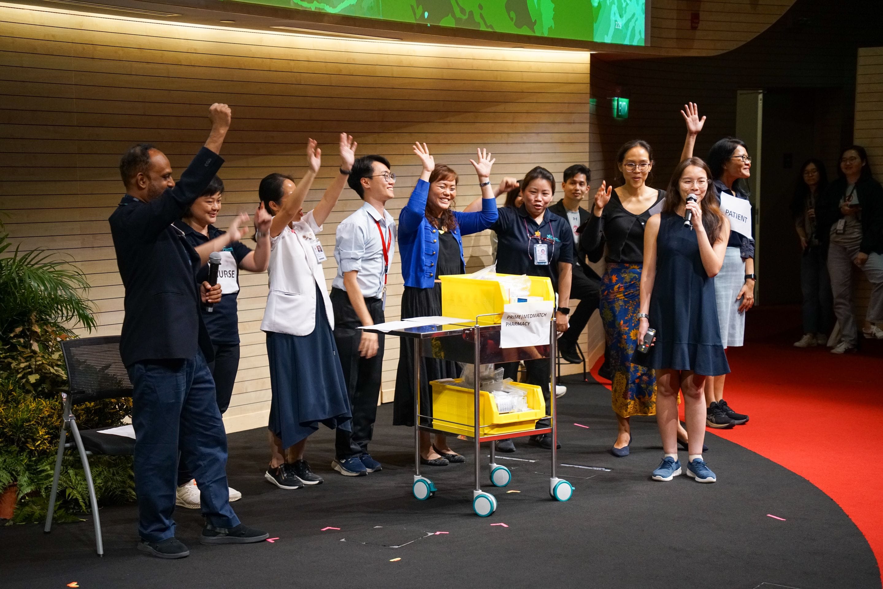 A group of people on a stage celebrating with raised hands near a yellow medical cart labeled "pharmacy."